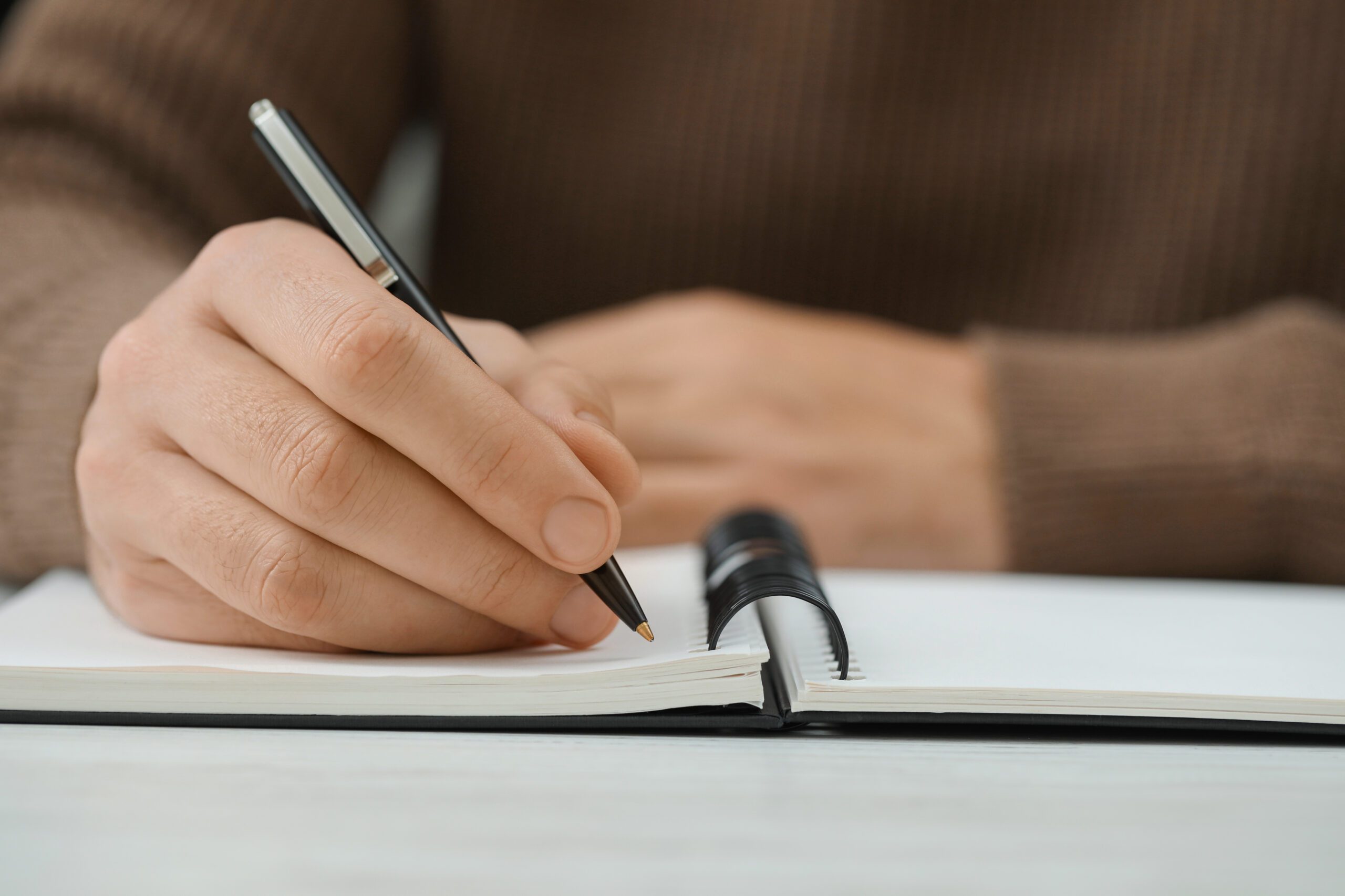 Man writing in notebook at white table, closeup. Space for text